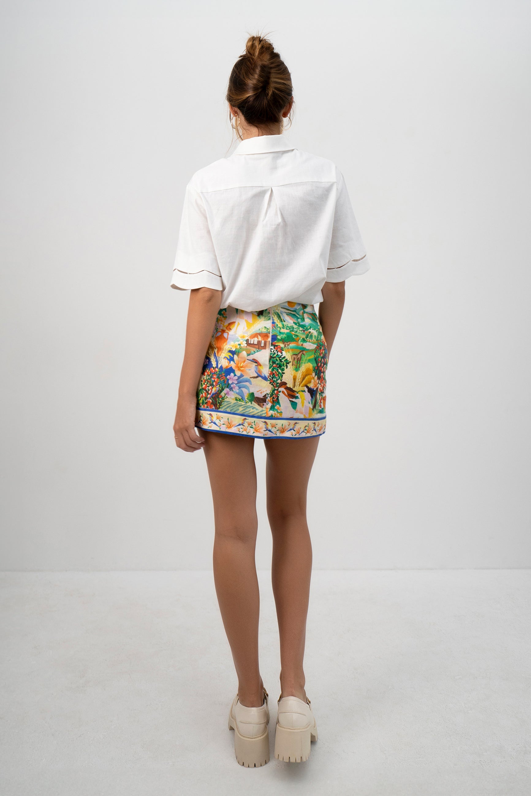 A model wearing the white Pilgrimage Shirt and the Halcyon Days Skirt asymmetrical faux wrap mini skirt featuring exclusive vibrant and picturesque textile print - back view shown.