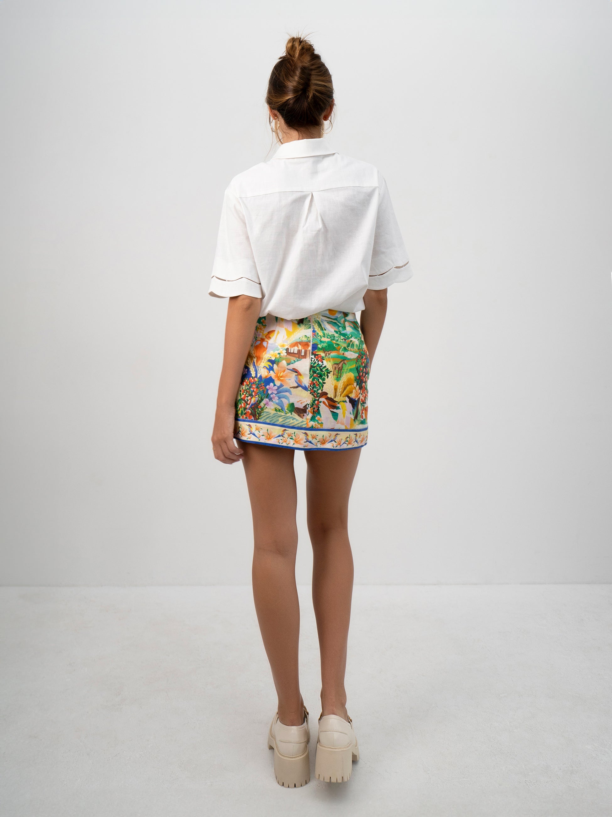 A model wearing the white Pilgrimage Shirt and the Halcyon Days Skirt asymmetrical faux wrap mini skirt featuring exclusive vibrant and picturesque textile print - back view shown.