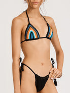 This image is a Front detail view of the INDIAN SUMMER BIKINI, a handcrafted swimwear piece from the Inherent Beauty collection. The model is showcased wearing the vibrant, multicolored top which is accented with black scallop-shaped edging, and black bottoms with tasseled side ties.